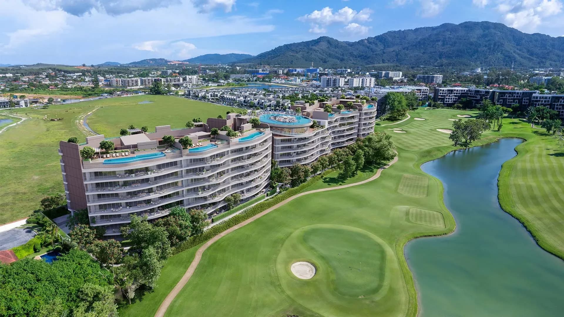 อาคารทรงโค้งสองหลังข้างกรีนและทะเลสาบของ Laguna Golf โดยมีภูเขาอยู่เบื้องหลัง
